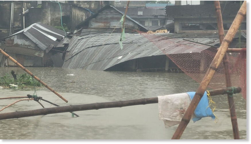 Tin-roofed houses partially submerged in floodwater in a low-lying area of Sylhet city.